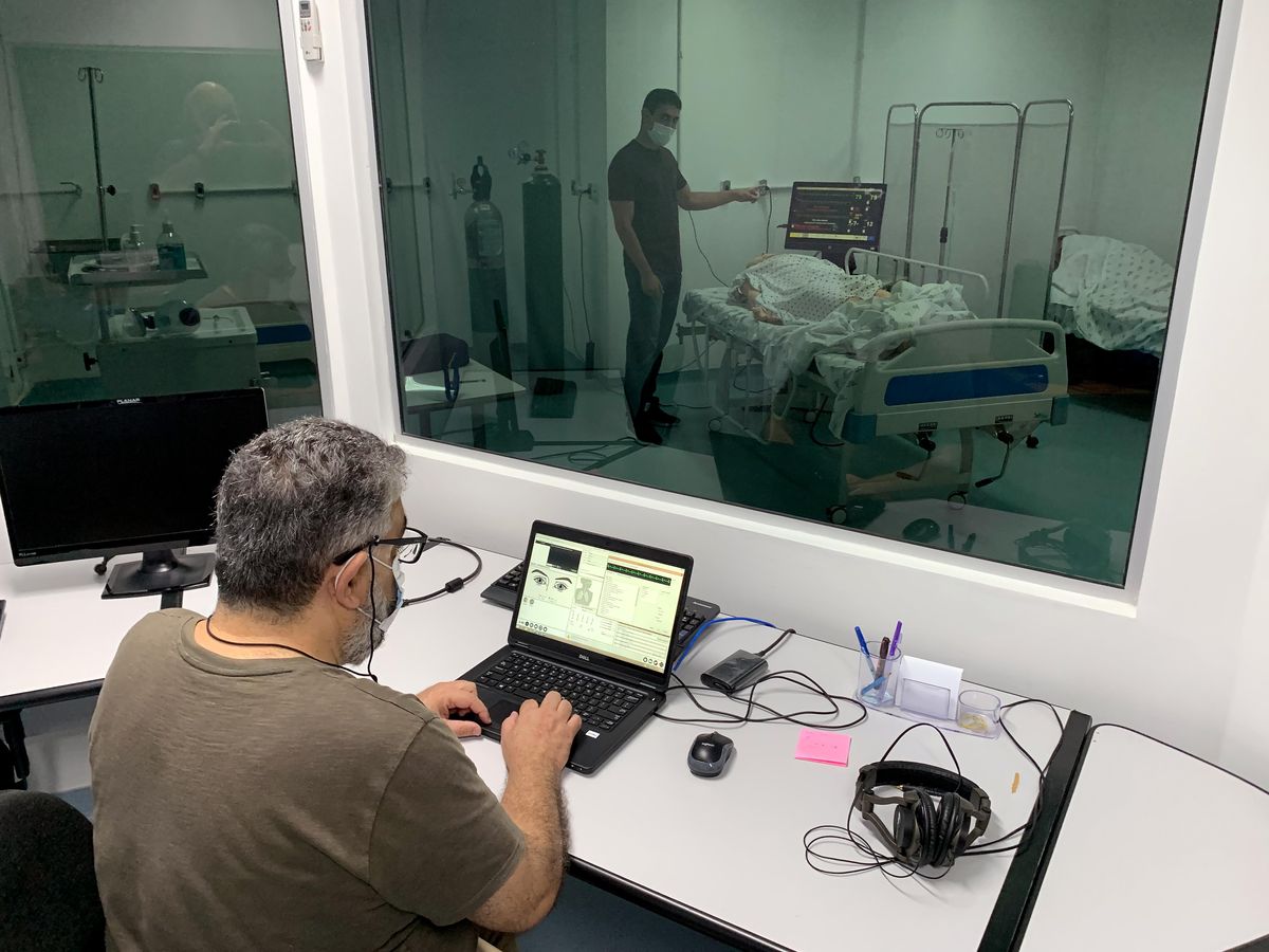 A middle-aged man monitors his younger colleague through a simulator lab window as he conducts heart-rhythm experiments on a medical dummy.